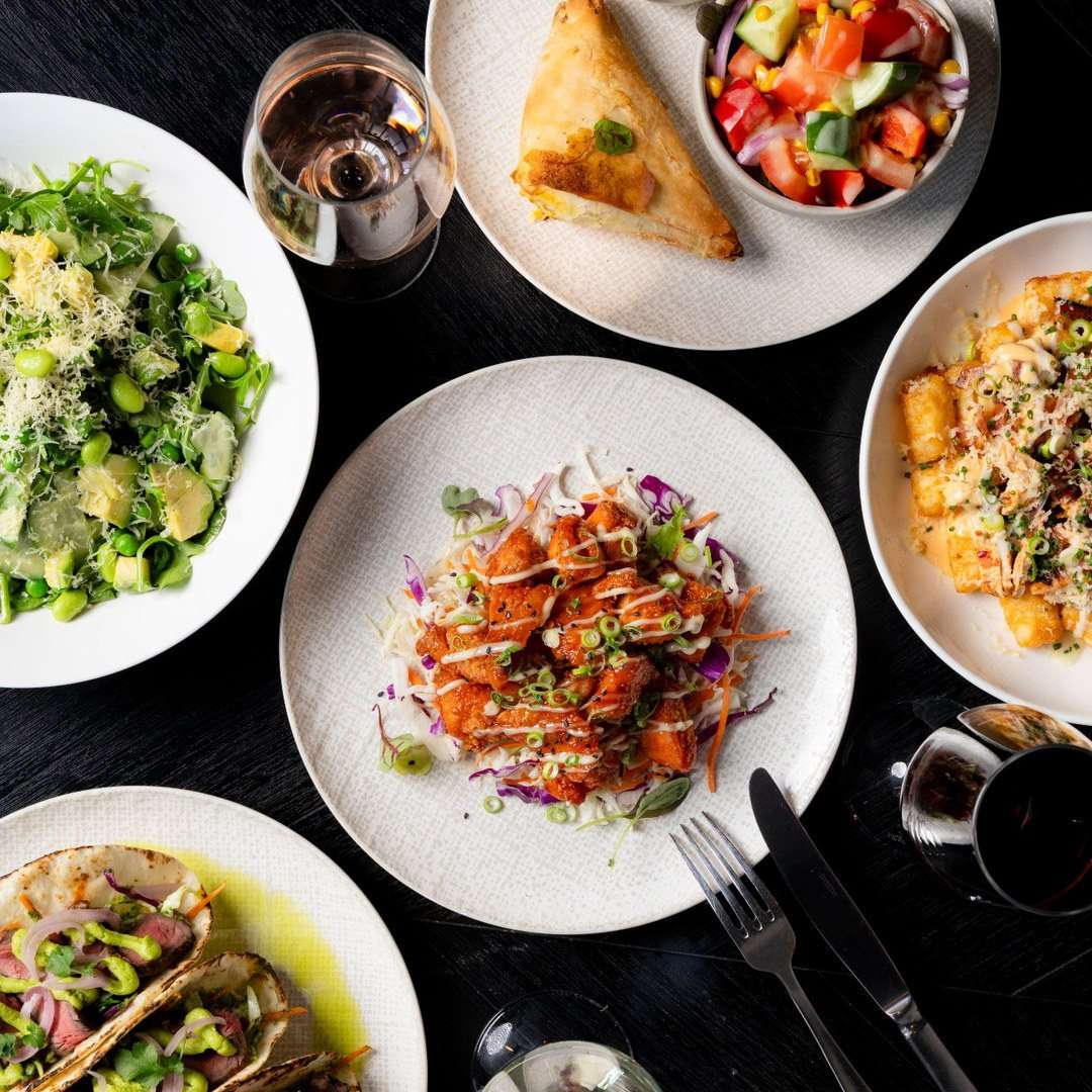 Overhead spread of spring menu dishes including tacos, salad, crispy bites and fresh sides with glasses of wine at Conservatory.